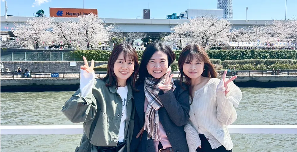 花見のイメージ写真。私服姿の女性社員3人が桜の木と河川敷をバックにして、ピースをして写っている。