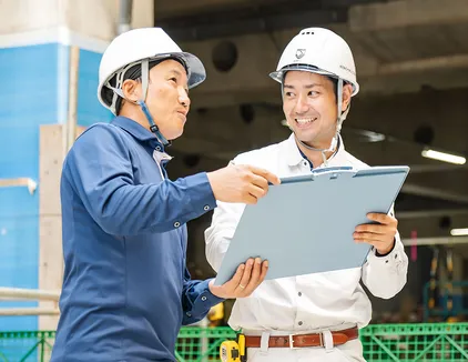 現場監督補助のイメージ写真。作業員2人がバインダーを見ながら話し合っている。